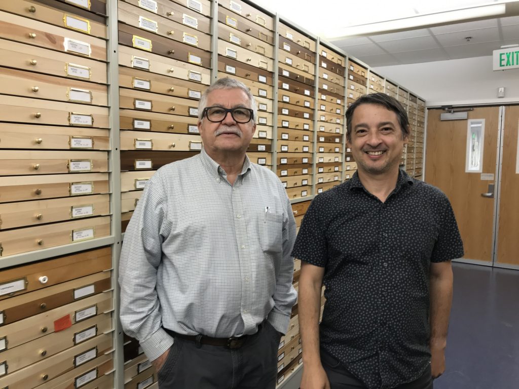 Two people posing for a photo in front of wooden specimen drawers