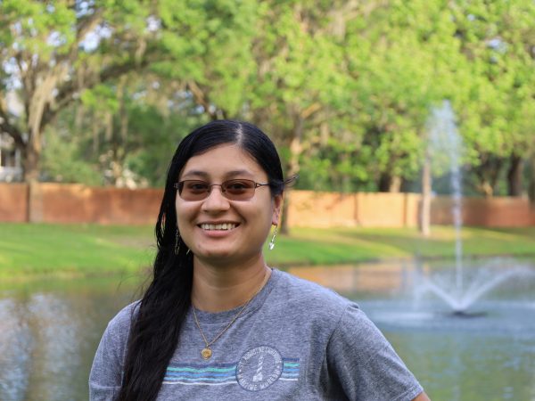 Woman wearing glasses stands in front of trees and fountain