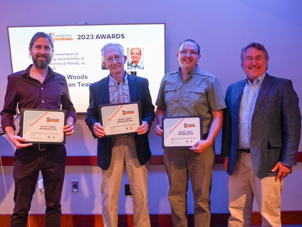 Four people stand in front of a screen, three of whom are holding award plaques