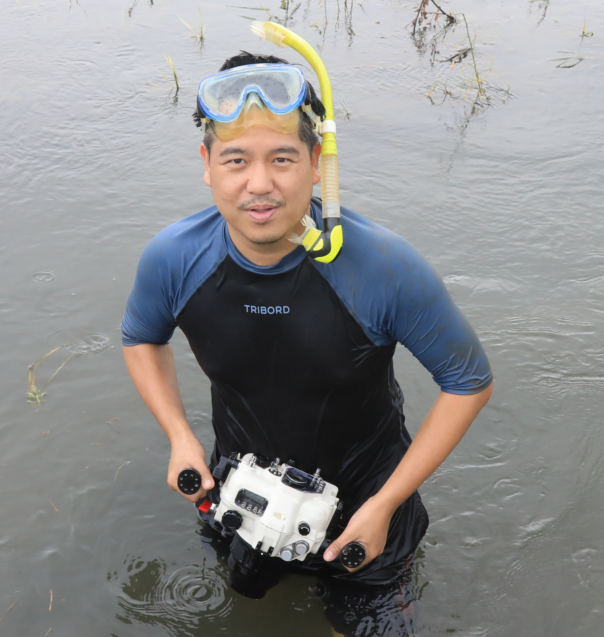 Man stands holding a waterproof camera while wearing a snorkel and googles in the middle of a river.