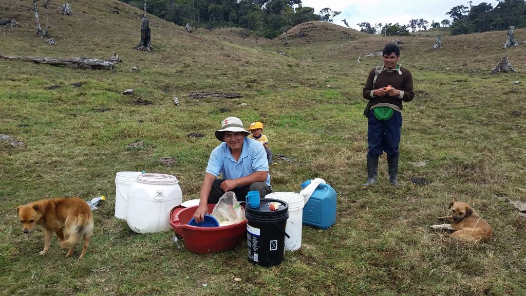 A man in a blue shirt and white hat smiles at the camera as he crouches over several buckets in a wide grassy field.