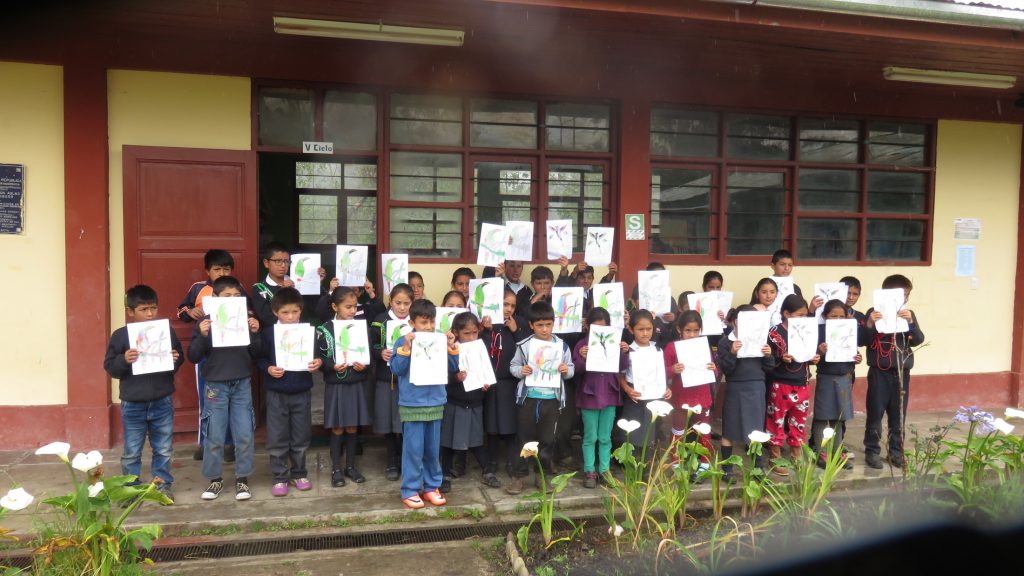 Dozens of children stand in a row outside a school building, each holding up a sheet of white paper with a hand drawn bird.