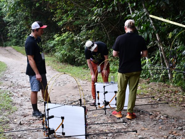 Three people stand on a gravel path assembling devices near aerial drones.