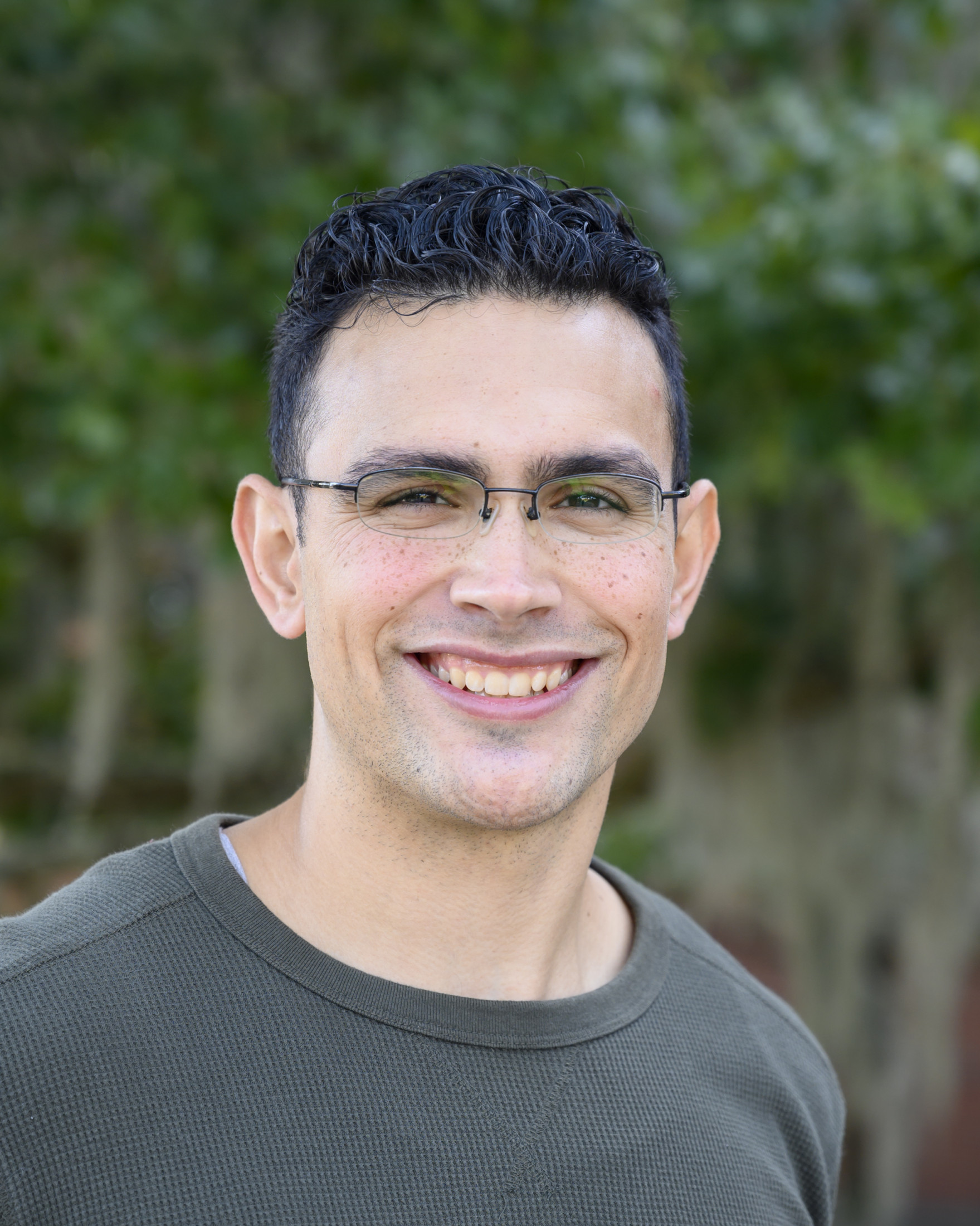 Person smiles at camera while standing in front of trees outside.