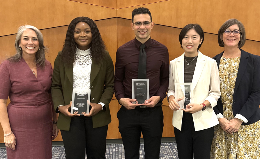 Five people smile for the camera, three of whom are holding award plaques. 