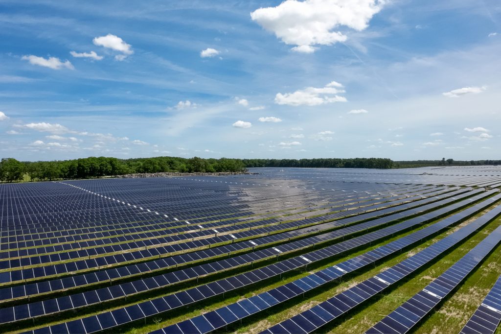 Photograph of solar arrays under a blue sky with trees and clouds in the background.