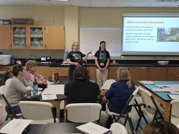 Two people stand at the front of a classroom next to a projection screen.