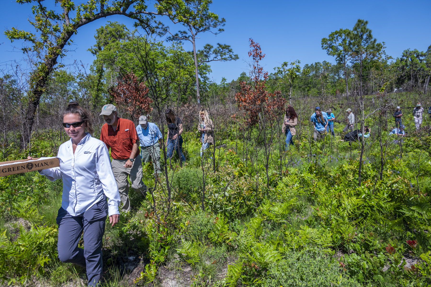 Several people walking in a line through a dry scrub environment under a clear blue sky.