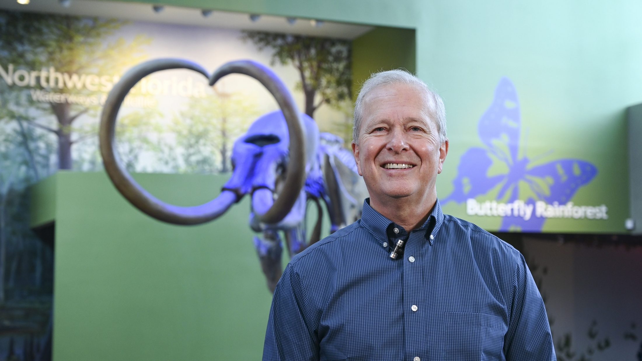Person smiles at camera in a natural history museum while standing in from of a mammoth. 