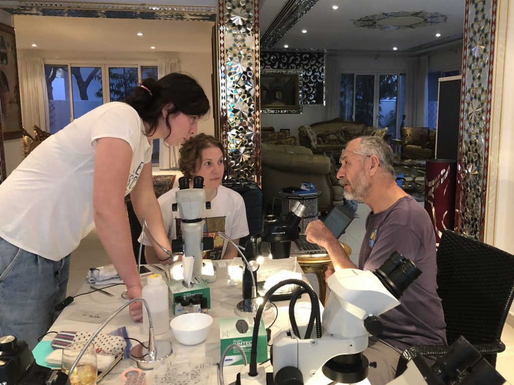 Photograph of three people sitting inside at decorative home. The table is full of lab equipment.