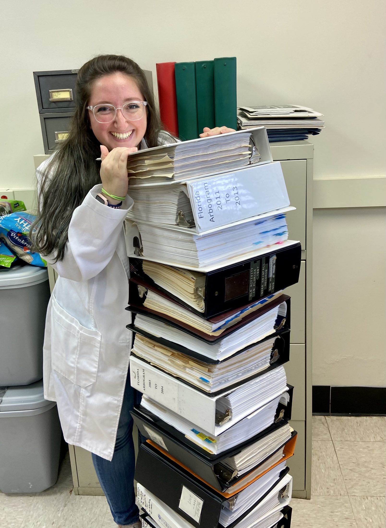 Person wearing a lab coat leans on a very tall stack of binders.
