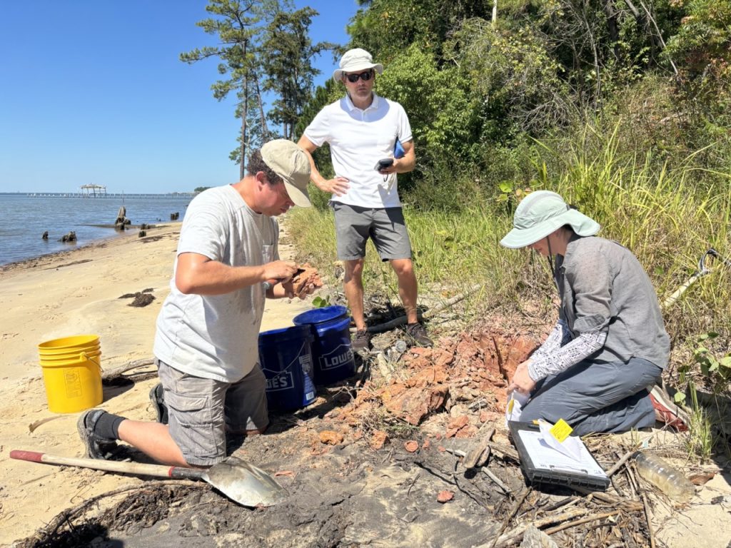 Photograph of three people kneeling on a beach with red clay, buckets, a shovel, and a clipboard around them.