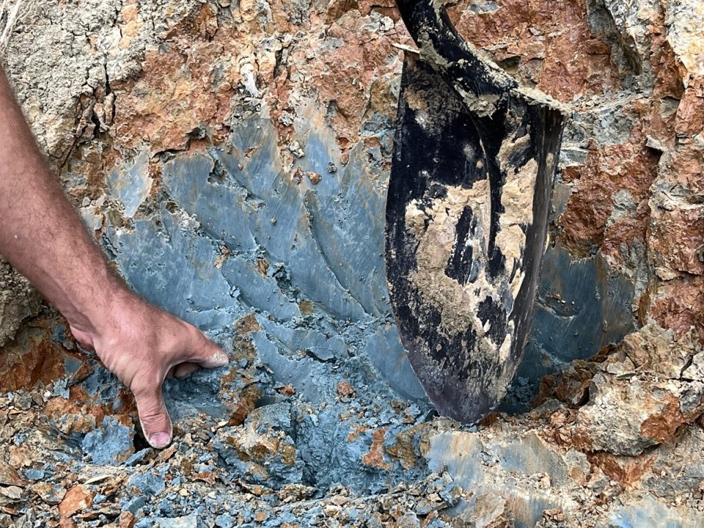 Photograph of a hand and a shovel digging into a bed of bright blue clay.