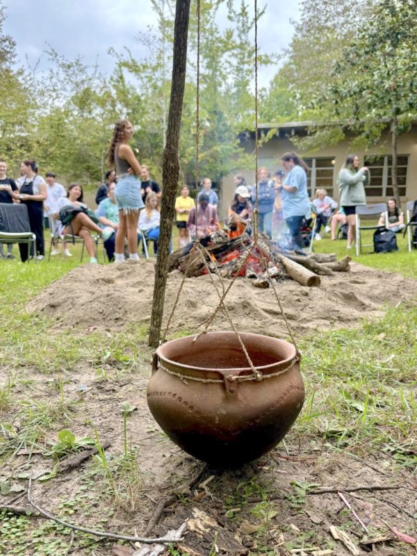 Photograph of a clay pot hanging by thin ropes with a bonfire and crowd of people in the background.