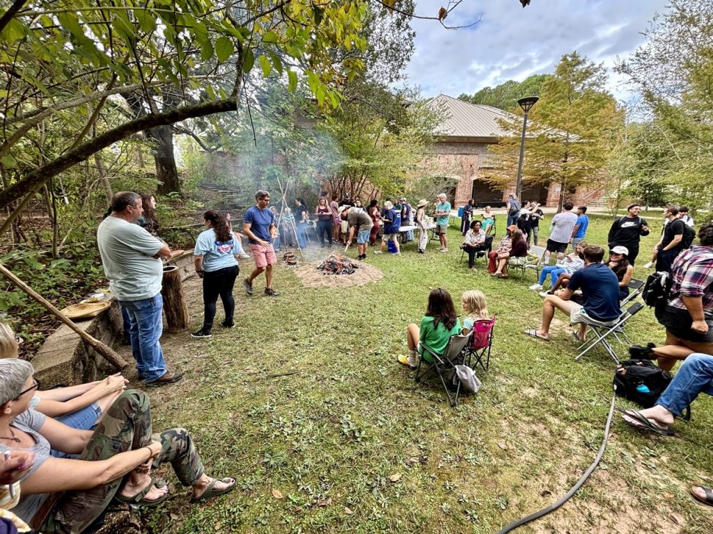Photograph of a crowd of people outside sitting around a smoking fire in the center.