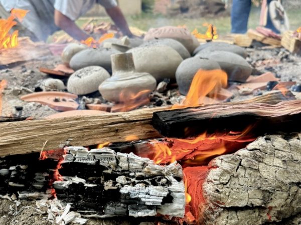 Photograph focused on a burning piece of wood, behind the wood is a pile of clay pottery and people bent over working.