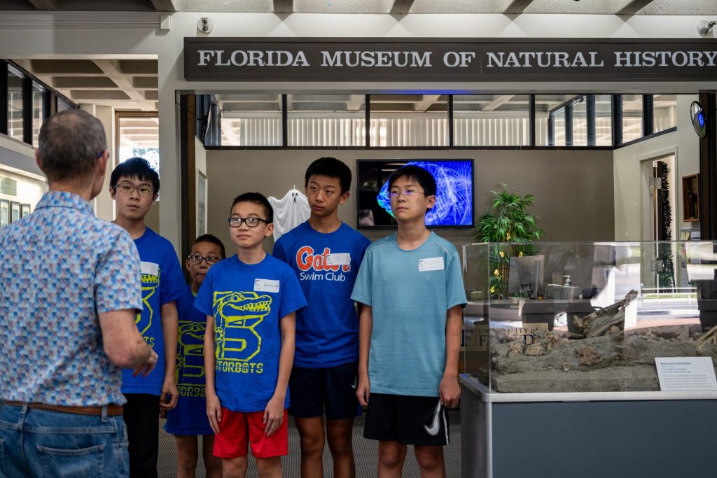 Photograph of a person addressing five kids standing in front of a Florida Museum of Natural History sign.