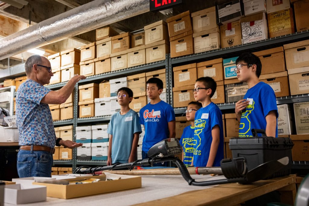 Photograph of a person talking to five kids standing in front of shelves of brown boxes.