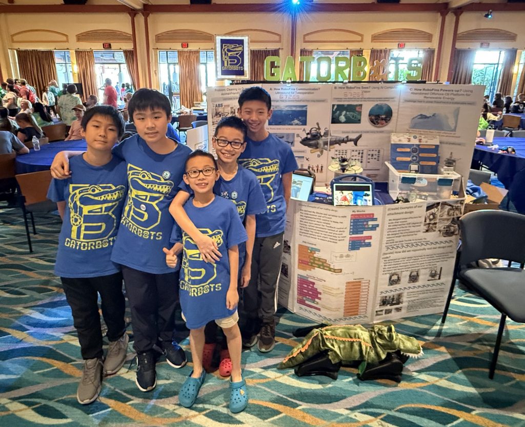 Photograph of five kids wearing blue team shirts and standing in an event space with a booth with posters of a research project.