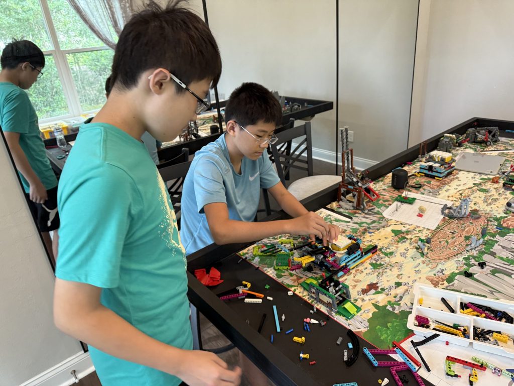 Photograph of two kids working on a robot with a pile of legos on the table in front of them.