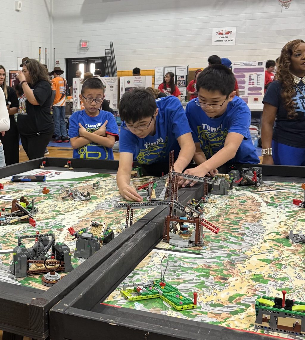 Photograph of three kids adjusting their robot while standing at a game table during a competition event.