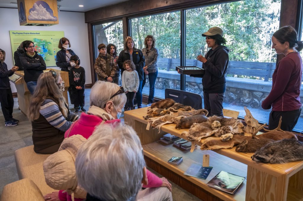two women standing behind a table of mammal skins and skulls, talking to a group of people