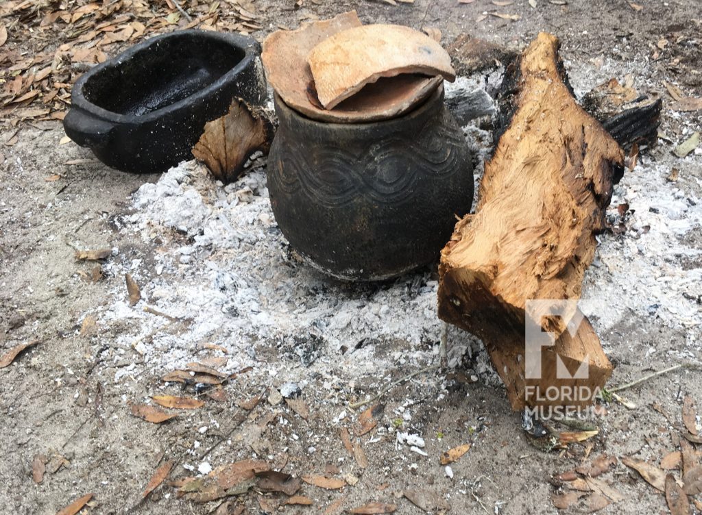 A blackened pot sitting in ashes with wood stacked next to it