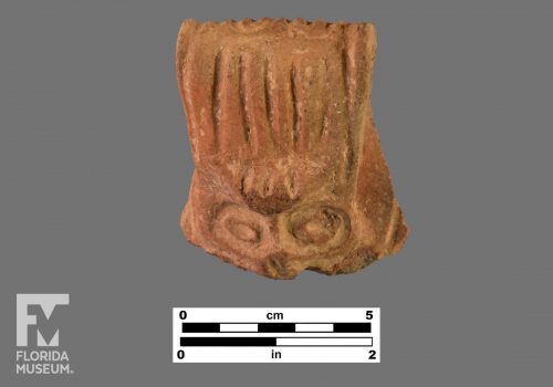 Formal artifact photo of an adorno face, with incised handle looped over to the back.