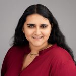 woman with dark hair wearing a red shirt standing in front of a grey background