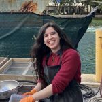 woman with long dark hair wearing a red shirt standing in front of a large boat