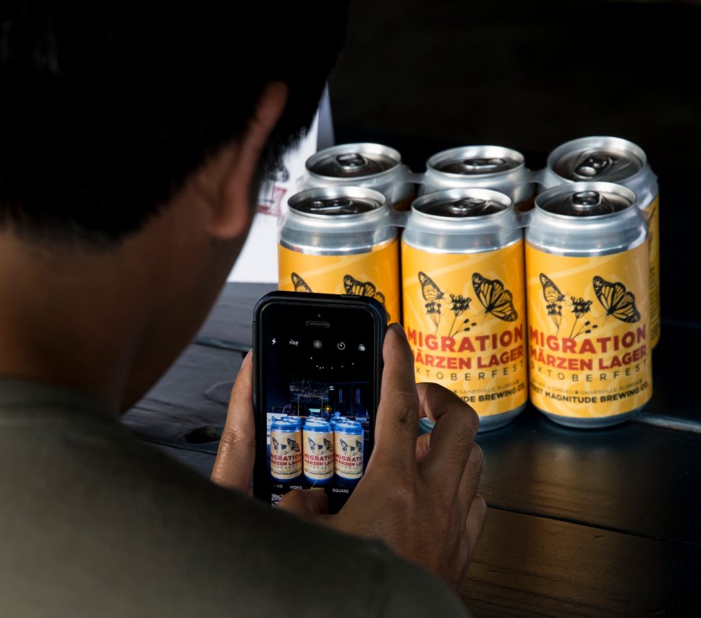 person using a phone to take a photo of a six-pack of the Migration Märzen Beer
