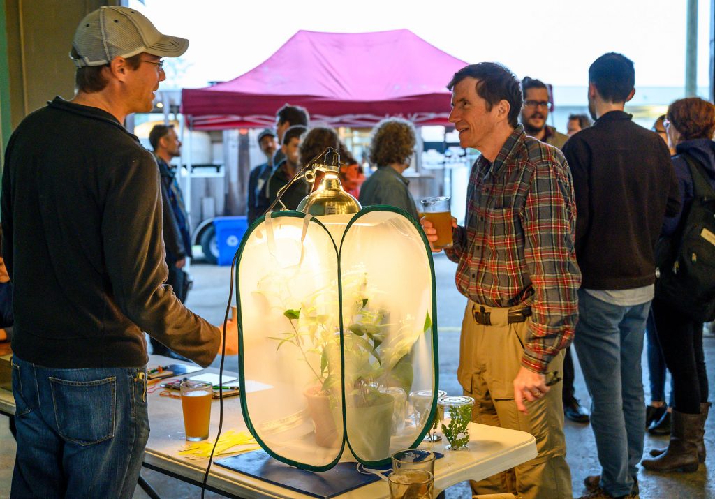 Volunteer Jim Grantham stands at a table with information about the Florida white butterfly and a terrarium with butterflies and speaks to a guest holding a beer.