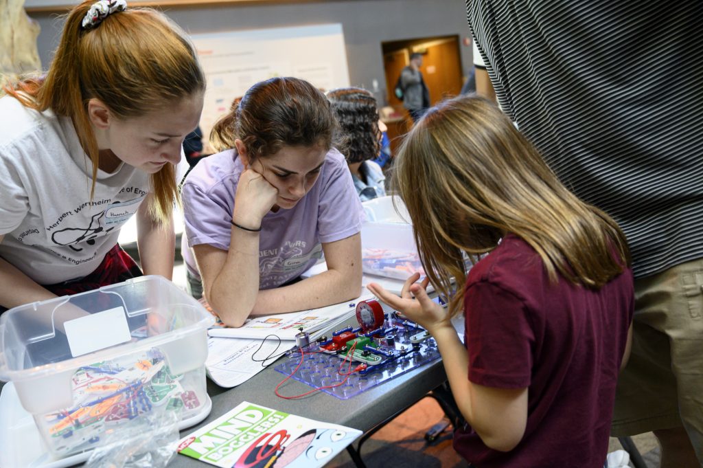 two students look on as a young girl works on a circuit board kit