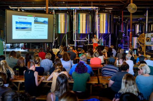 woman in front of large brewery vats talking to an audience