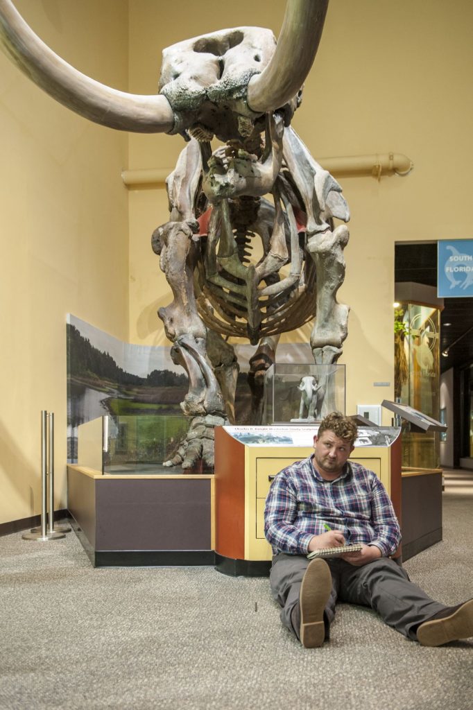 artist sitting on the floor in front of a mastodon fossil while drawing