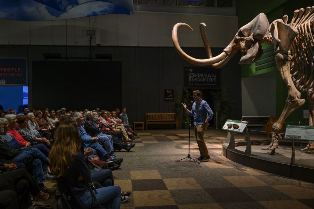 Randy Singer stands in front of mammoth skeleton and speaks to audience at the Fieldwork Fails with Guts & Glory GNV event.
