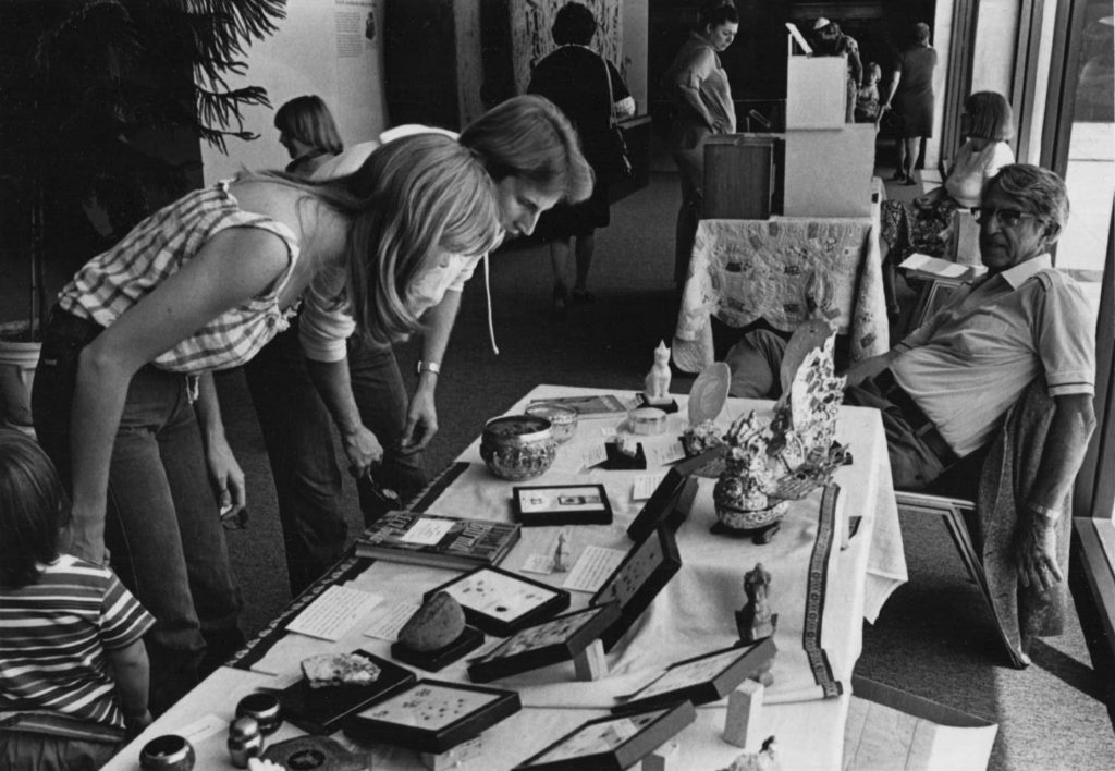 Black and white photo of people looking at an assortment of objects on a table.