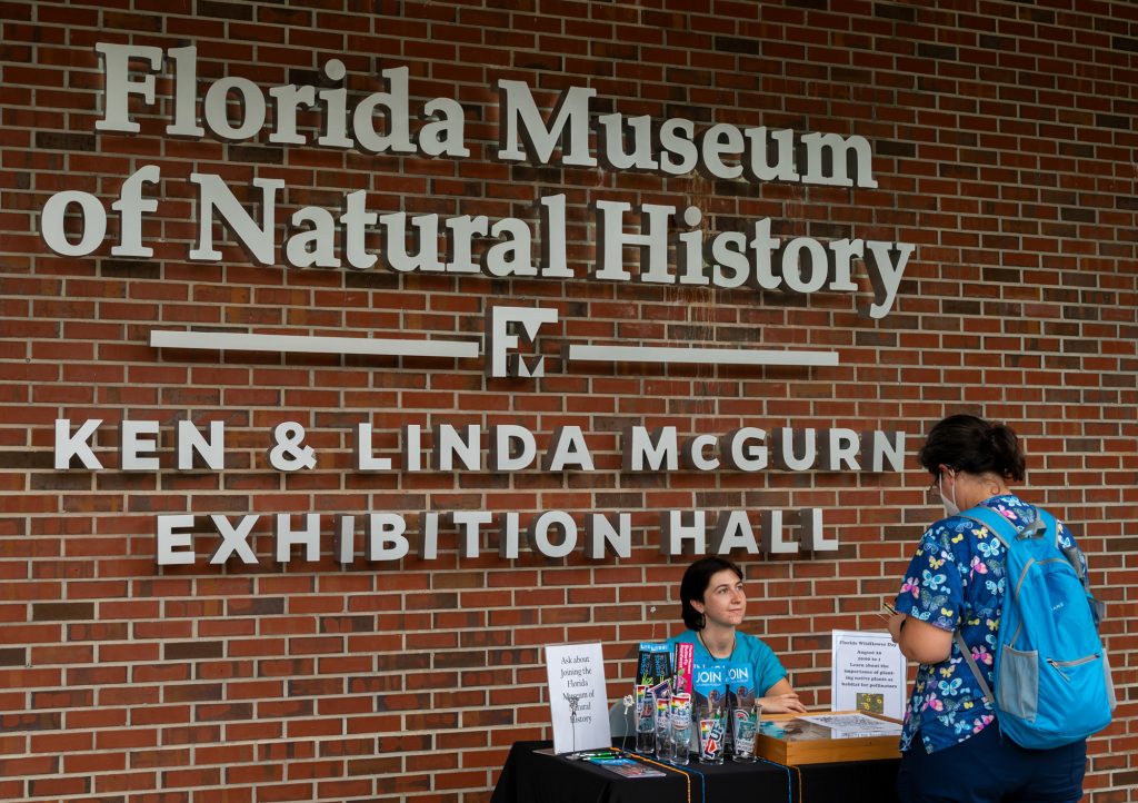 museum worker sits at a display table in front of the Florida Museum sign and speaks with visitor