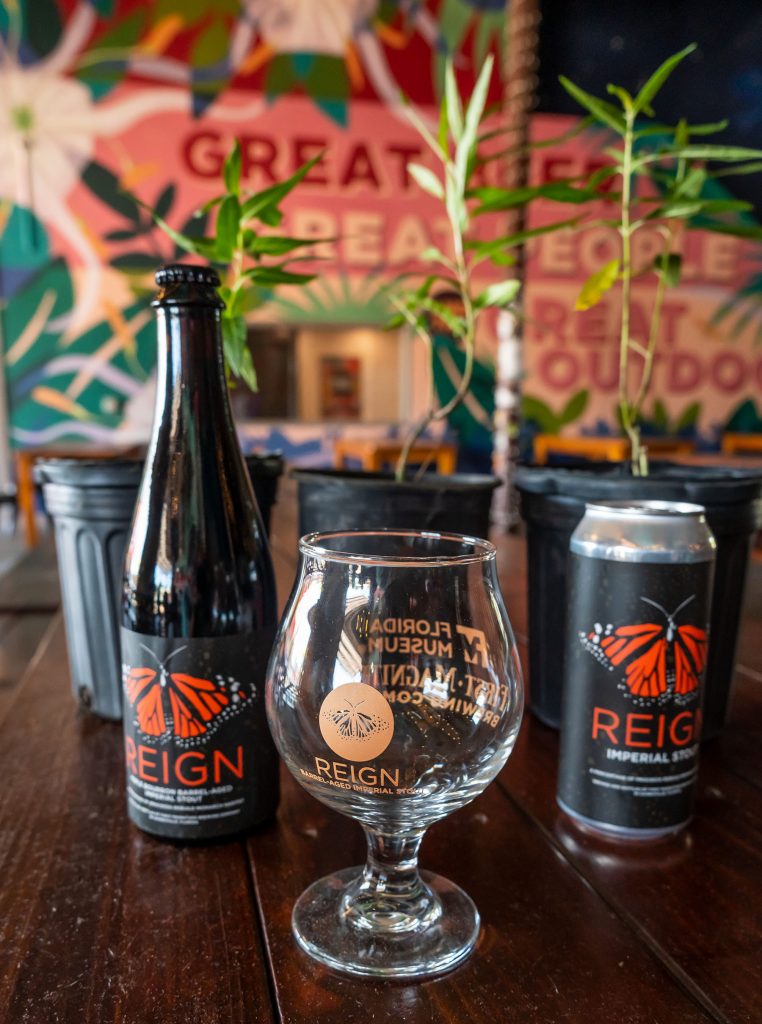 Bottled and canned imperial stout each with the Reign and orange butterfly logo sit next the Reign tulip glass. Behind the bottle, can, and glass are potted milkweed plants and the brightly colored murals at First Magnitude brewery