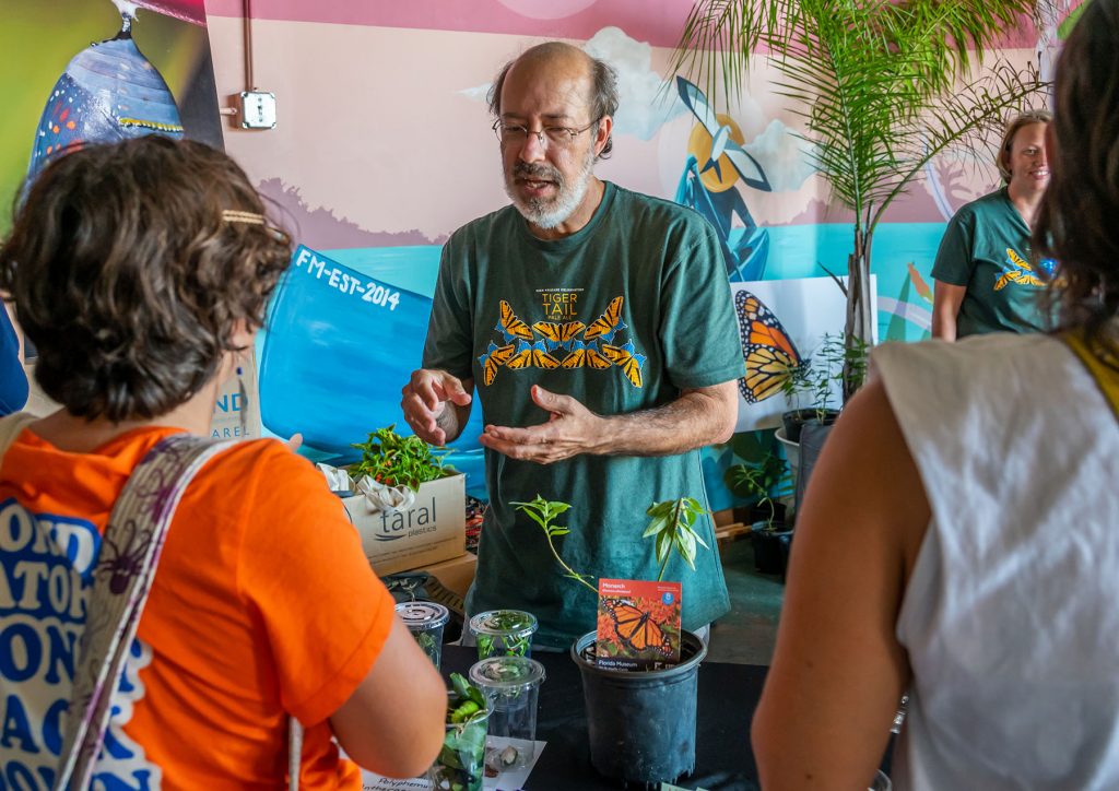 Jaret Daniels stands behind a display table willed with milkweed plants and butterfly specimens and speaks with visitors during the beer launch