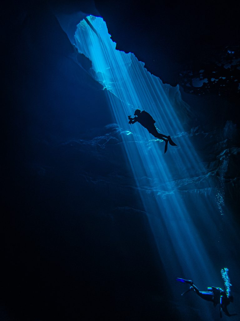 Two divers swim in shafts of light shining through the entrance of the underwater cave in Buford Sink