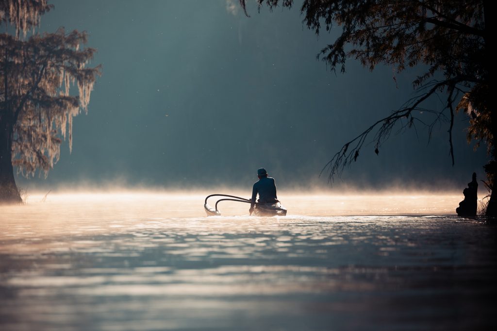 A person in a small boat sits at the center of the river silhouetted against the mist rising off the water