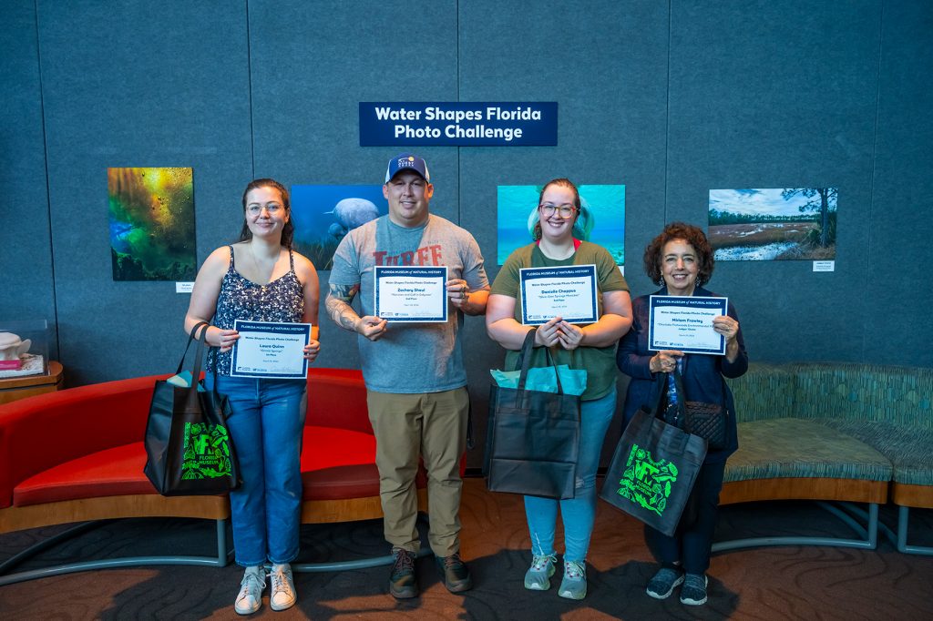 four people holding printed certificates stand in a museum gallery in front of printed photos of under water scenes