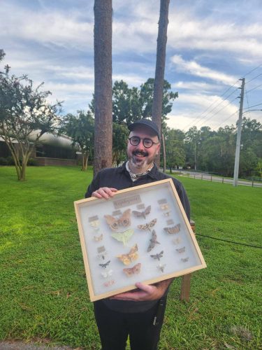 man, Vaughn Shirey, standing outside holding a specimen container with many variety of pinned moth specimens 