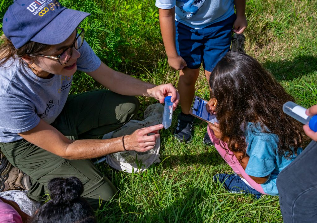 Kaitlin Deutsch holding a collection tube with a captured insect while a child writes observations using a clipboard.