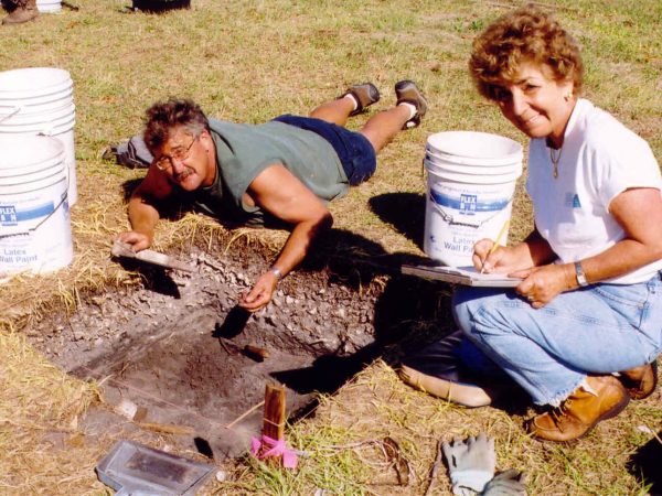 one person laying on the ground looking in s shallow excavation pin, other person kneeling next to the pit taking notes on a clipboard