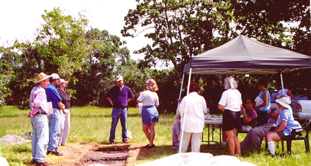 group of researchers standing in a field, some under a tent listening to one of the researchers speak