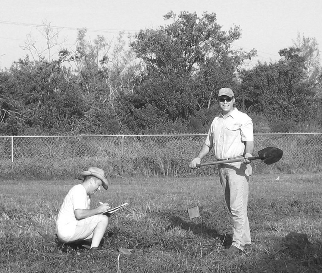 man stands with a shovel while another man kneels on the ground and takes notes in a notebook