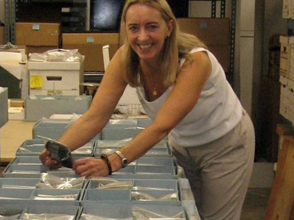 person leaning over a table filled with small blue boxes containing archaeobotanical samples in plastic bags. She is looking and smiling at the camera
