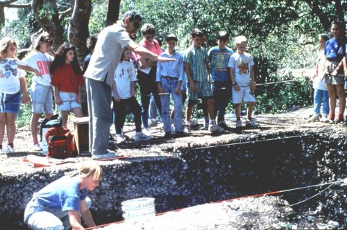 children and adults stand at the edge of an archeological pit, One person gestures into the pit. 